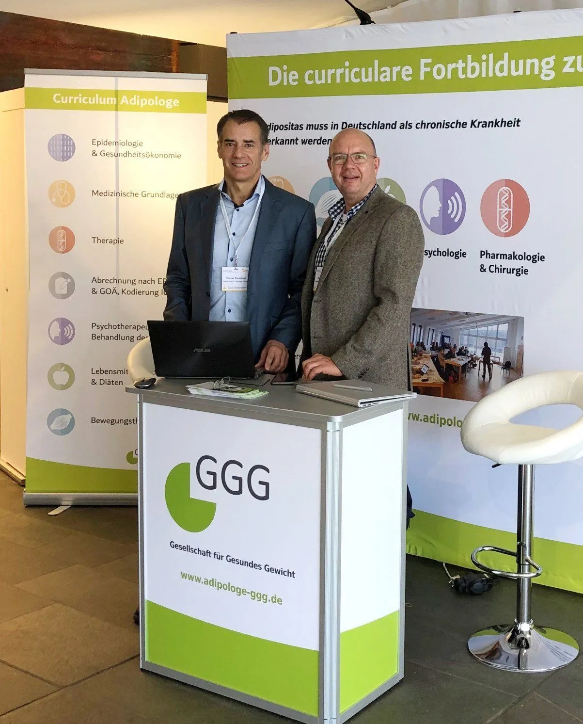 Zwei Männer am Ausstellerstand der GGG mit Stehtisch, GGG-Logo und Roll-ups zum Curriculum Adipologe