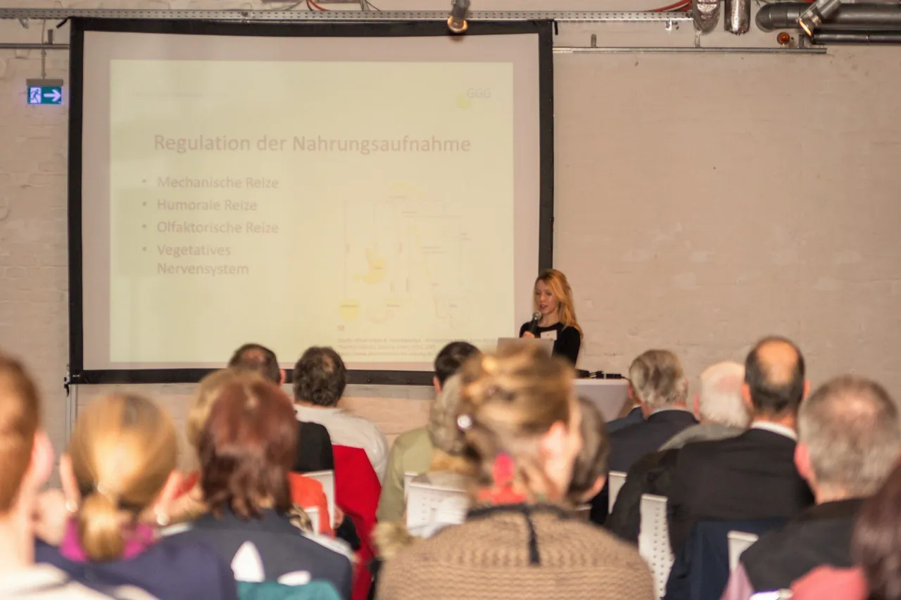 Eine Referentin mit Mikrofon vor der Leinwand mit der Folie „Regulation der Nahrungsaufnahme“, im Vordergrund das Publikum