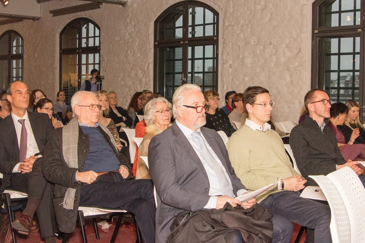 Zuhörerinnen und Zuhörer in Stuhlreihen vor hohen Rundbogenfenstern im Veranstaltungssaal